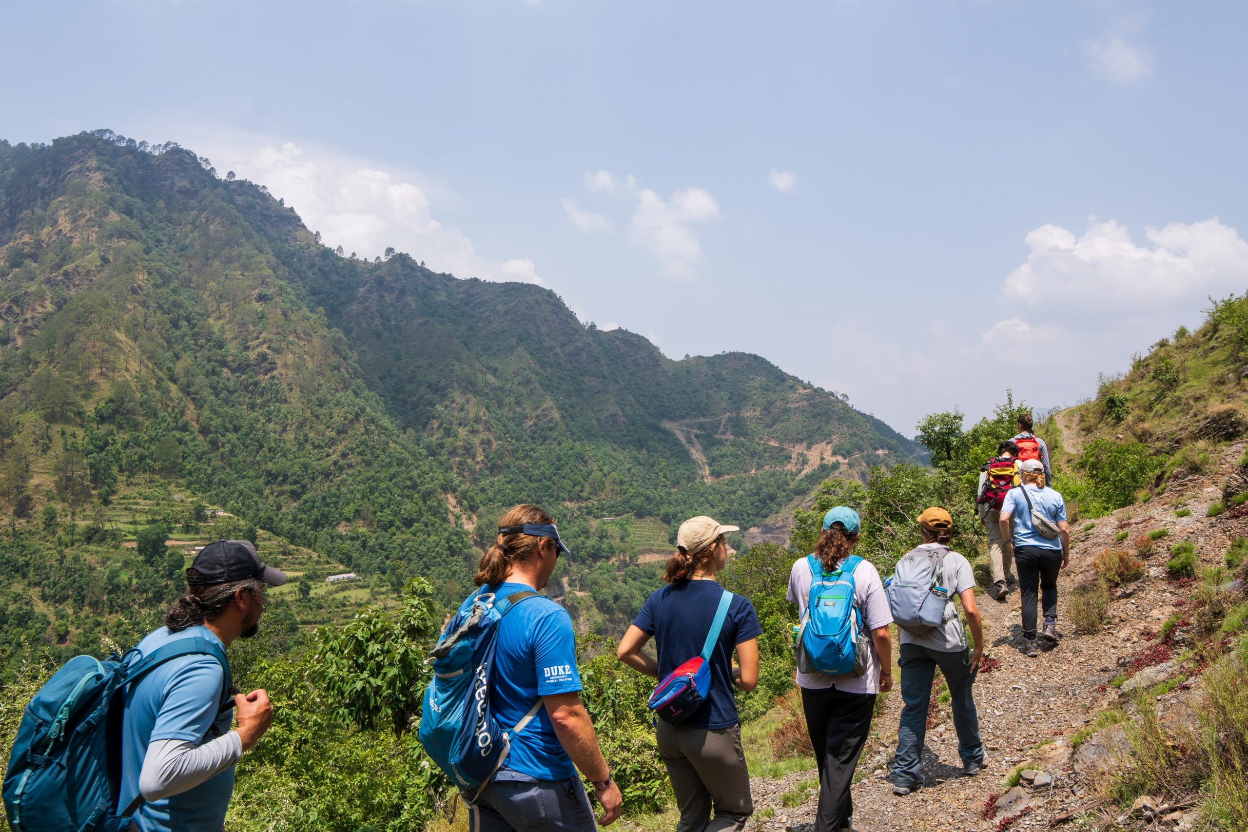 A group of people wearing short-sleeved shirts, pants and backpacks and baseball hats hike up a gravelly mountain in a single line.