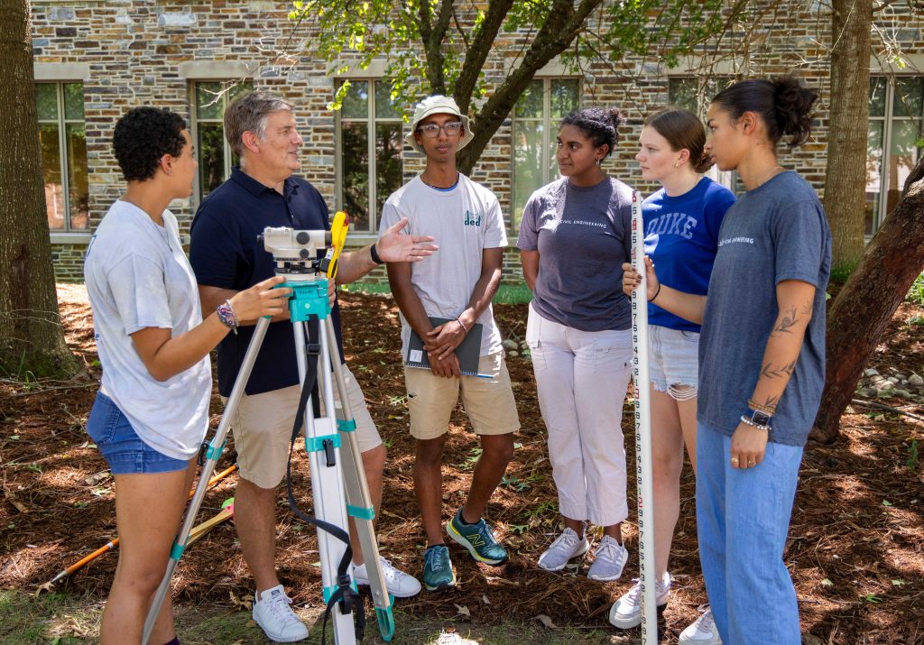 Man talking with a group of students. One student is holding a surveyor camera on a tripod and the other is holding a measuring stick.