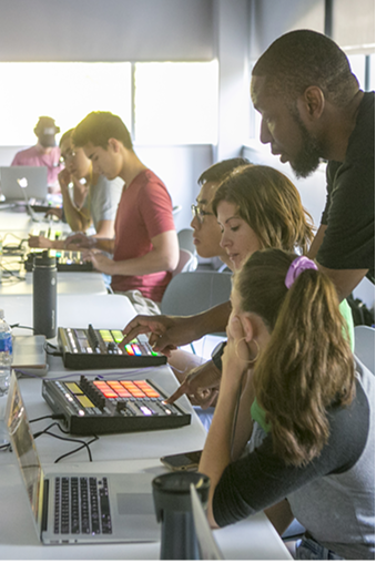 Profile view of several students sitting at a table working on computer equipment and laptops.