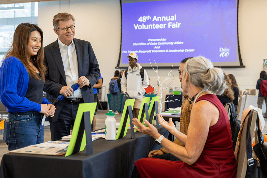 A female student in a blue cardigan and jeans stands with Duke president in a navy suit and glasses, talking to two people sitting at long table with a black tablecloth and green tabletop signs propped up. One woman sitting has a red sleeveless dress on.