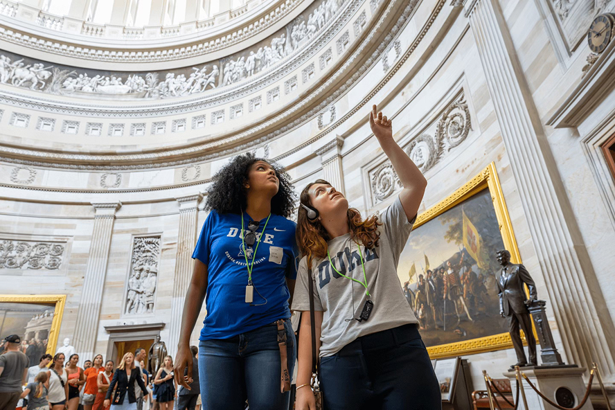Two female students wearing Duke t-shirts pointing at the ceiling of the Capitol building in DC, surrounded by paintings and statues of the founding fathers.