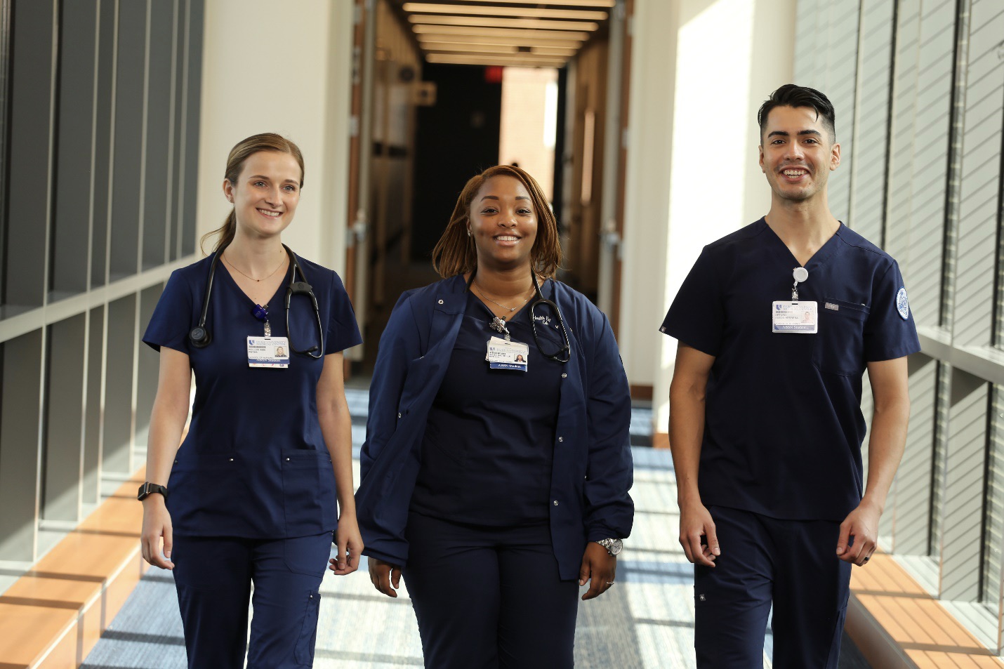 Three nursing students standing in a sunny hallway with windows on both sides